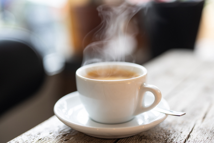 Steam rising from a white cup of hot coffee with a spoon on a saucer over a wooden table in the cafe.